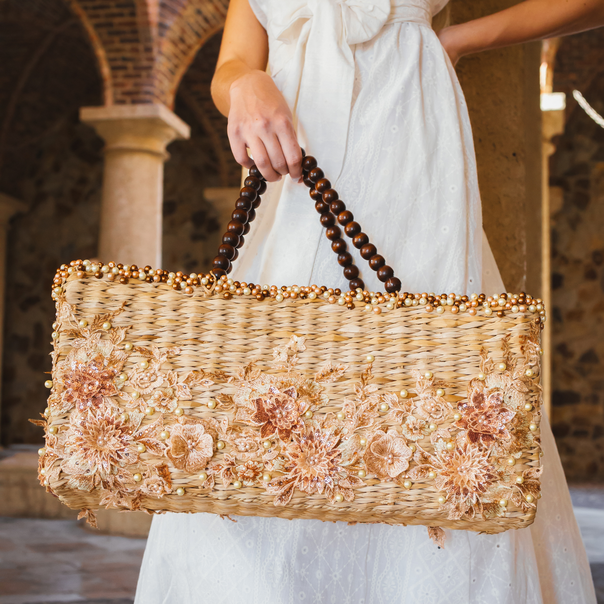 Woman holding a rectangular wicker handbag with rose appliqués, pearl accents, and dark wooden bead handles, styled with a white dress in a rustic courtyard