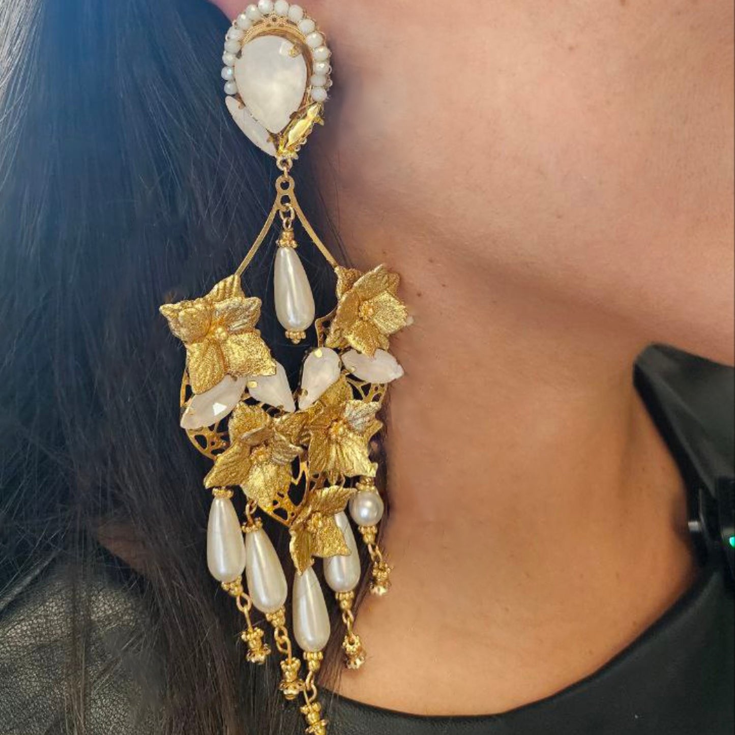 Close-up of a woman wearing a long gold floral earring with pearl and crystal details, styled with dark hair and a black top.