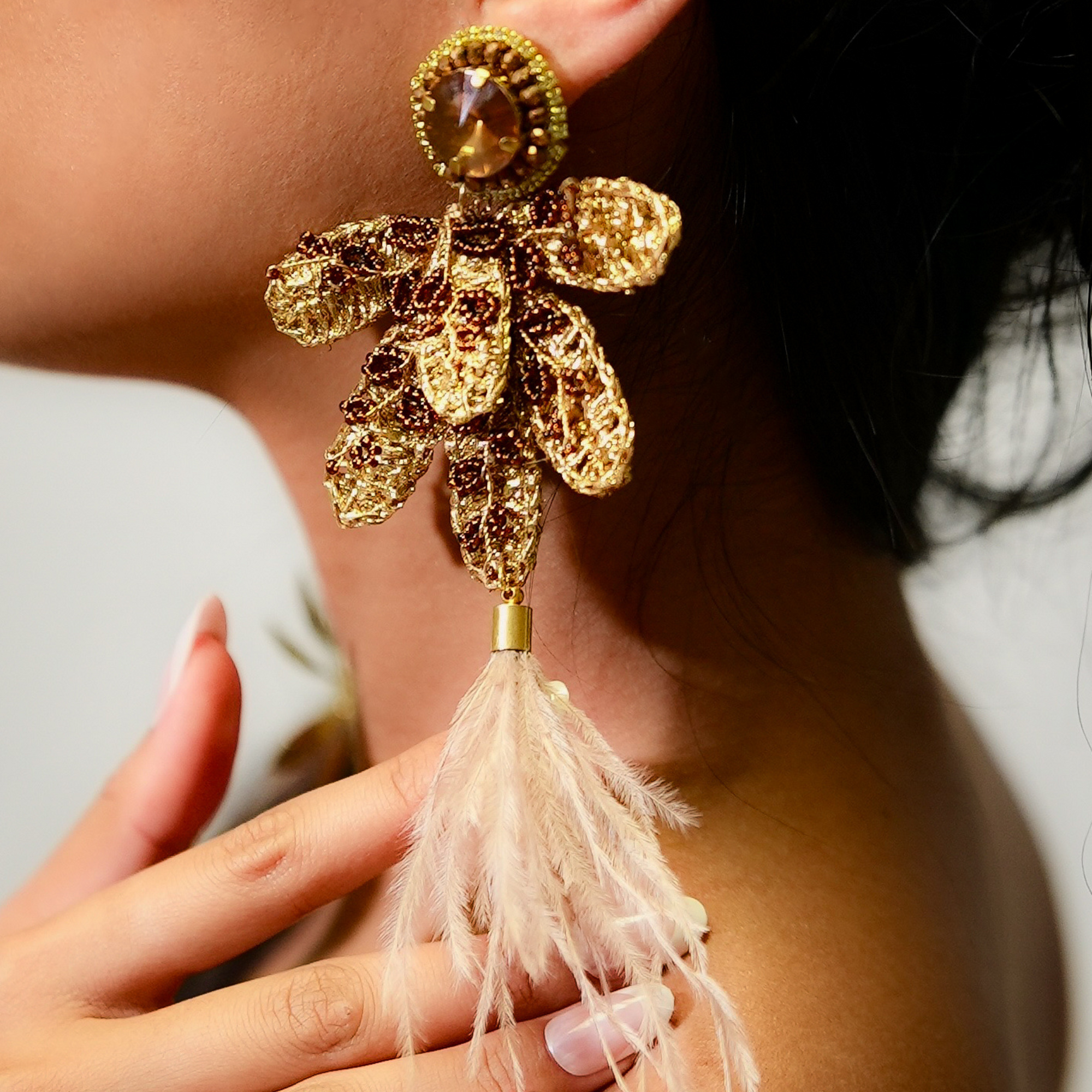 Gold and brown feathered earring held by a hand against a neutral background