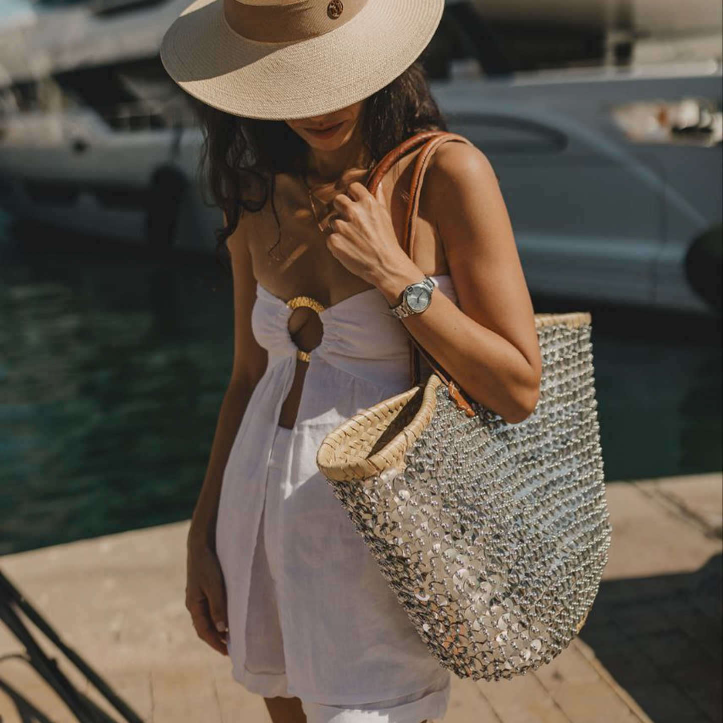 Woman carrying the Silver Lentejuelón Bag in a coastal setting, wearing a white summer outfit and sunhat