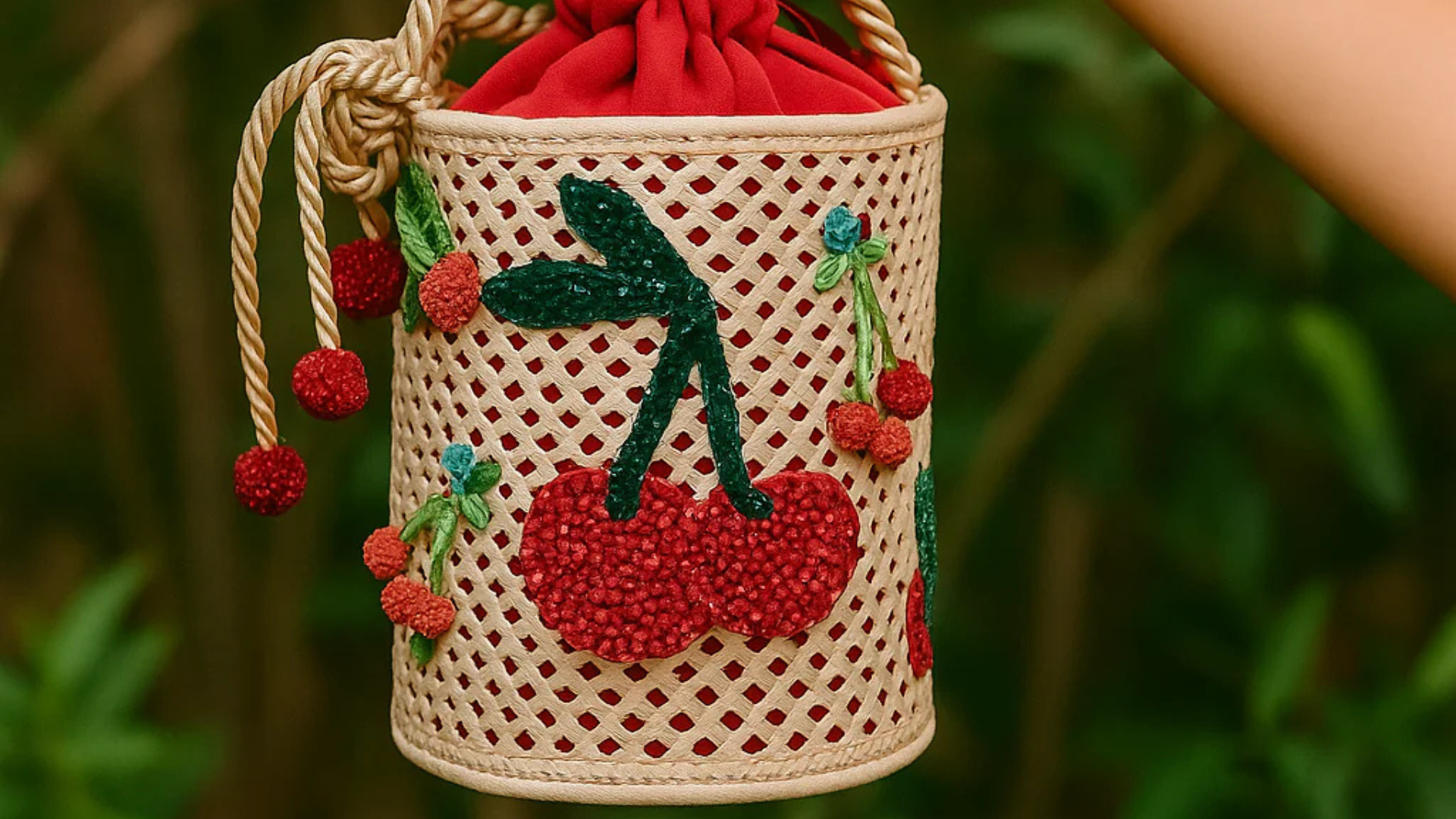Close-up of a cherry-themed cylindrical woven handbag with embroidered red cherries, green leaves, and red fabric lining, held outdoors against a green background.