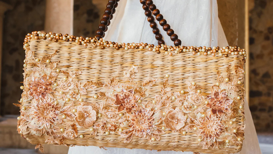 Woman in white dress holding a rectangular woven handbag adorned with blush floral embroidery, pearl accents, and dark wooden beaded handles.