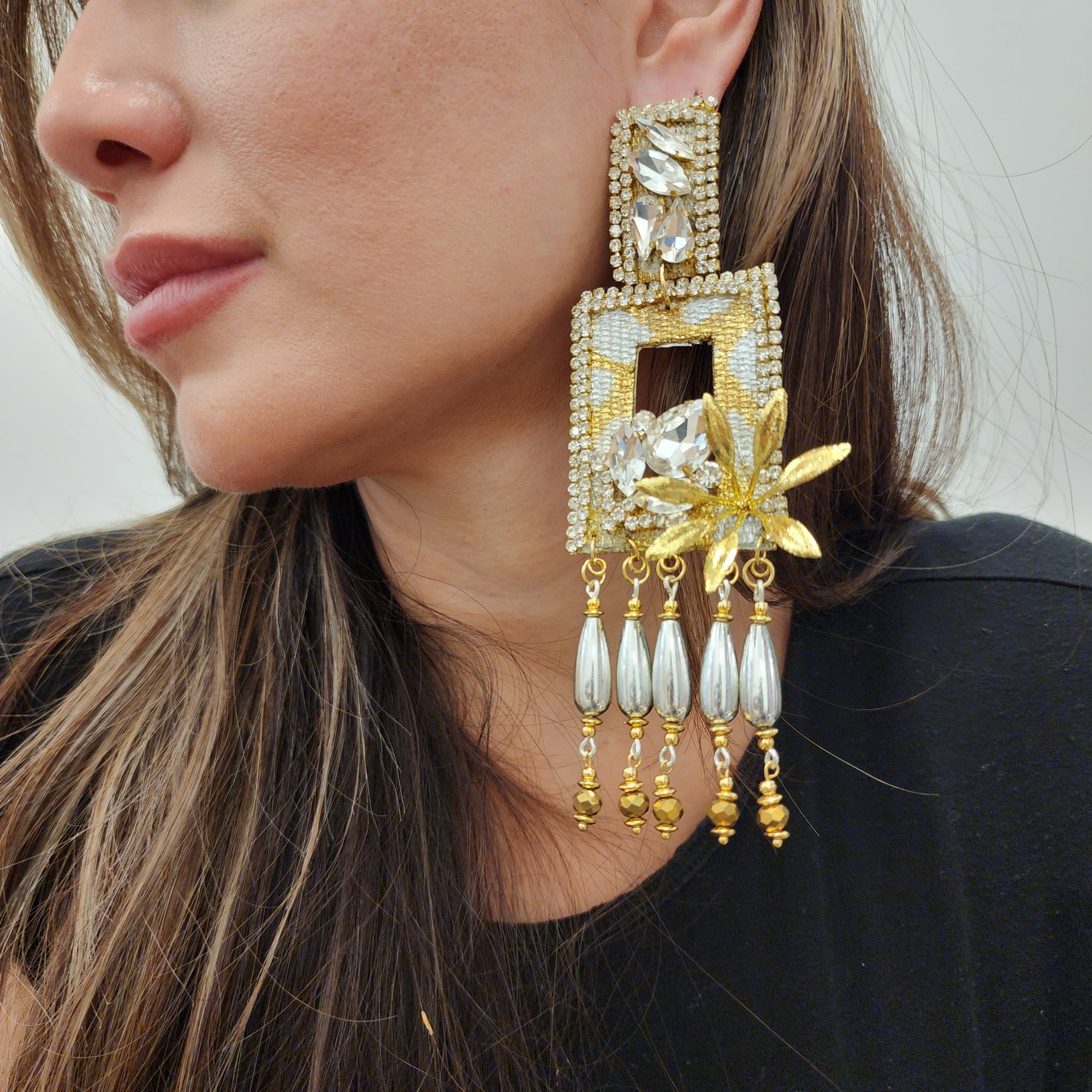 Close-up of a woman wearing a large rectangular gold and crystal statement earring with floral detail and dangling pearl-like beads.