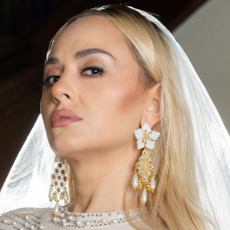 These beautiful earrings feature intricate gold designs, crystal embellishments, and dangling pearl-like beads. A delicate white beaded floral motif at the top enhances the elegance of the design, making them perfect for formal or festive occasions.