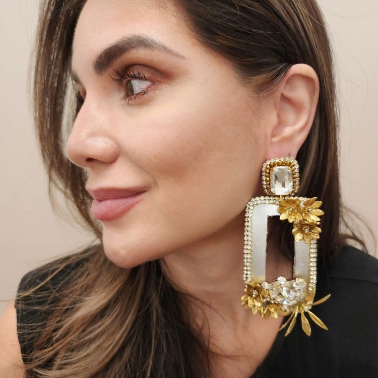Close-up of a woman wearing a large rectangular gold and crystal earring adorned with floral embellishments and rhinestone accents.