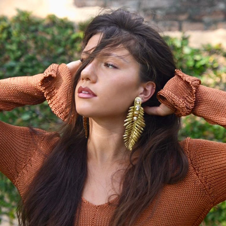 Woman wearing a brown sweater and gold earrings outdoors with greenery and stone steps in the background