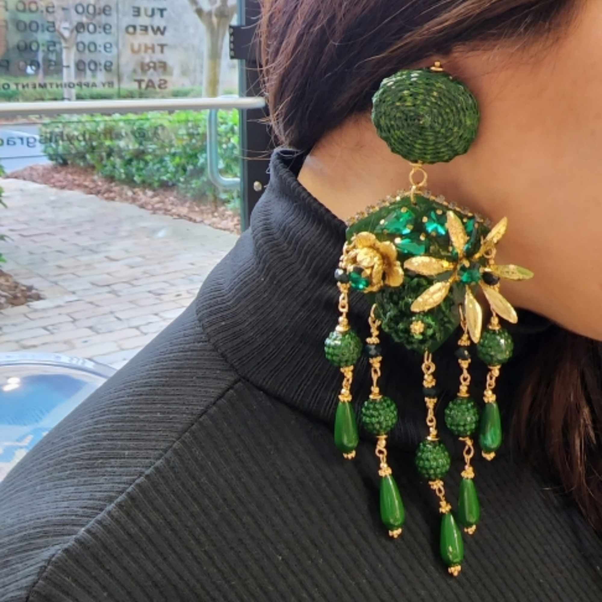 Close-up of handcrafted green earrings with gold floral accents and beaded drops, worn with a black textured top for a bold contrast.