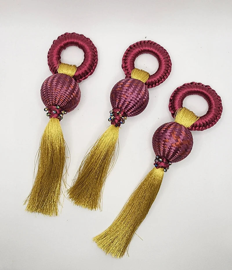 Each napkin ring features a circular design at the top, followed by a purple-hued woven sphere and a gold fringe hanging from each