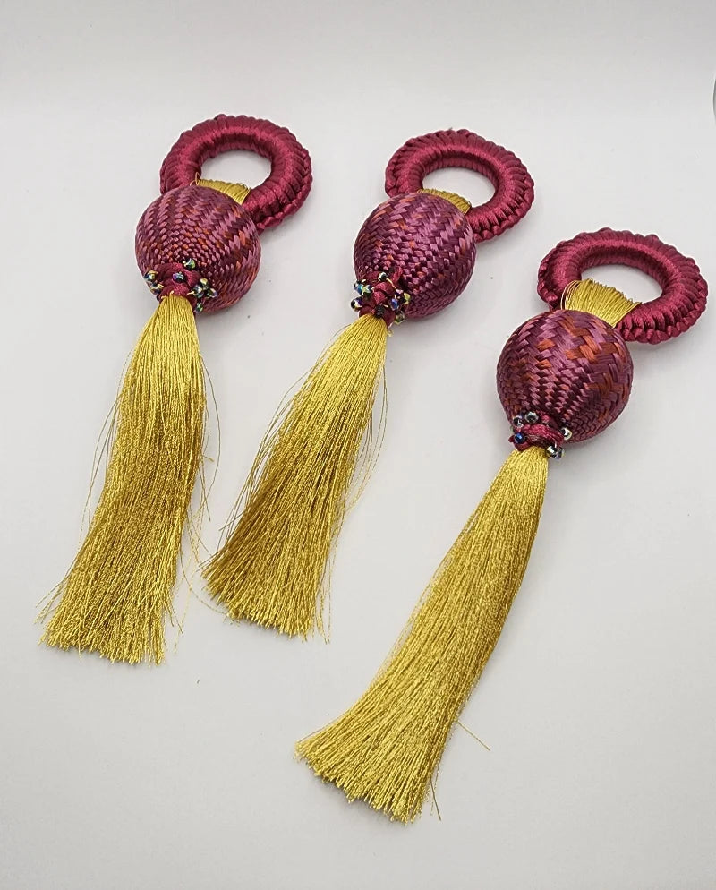 Each napkin ring features a circular design at the top, followed by a purple-hued woven sphere and a gold fringe hanging from each