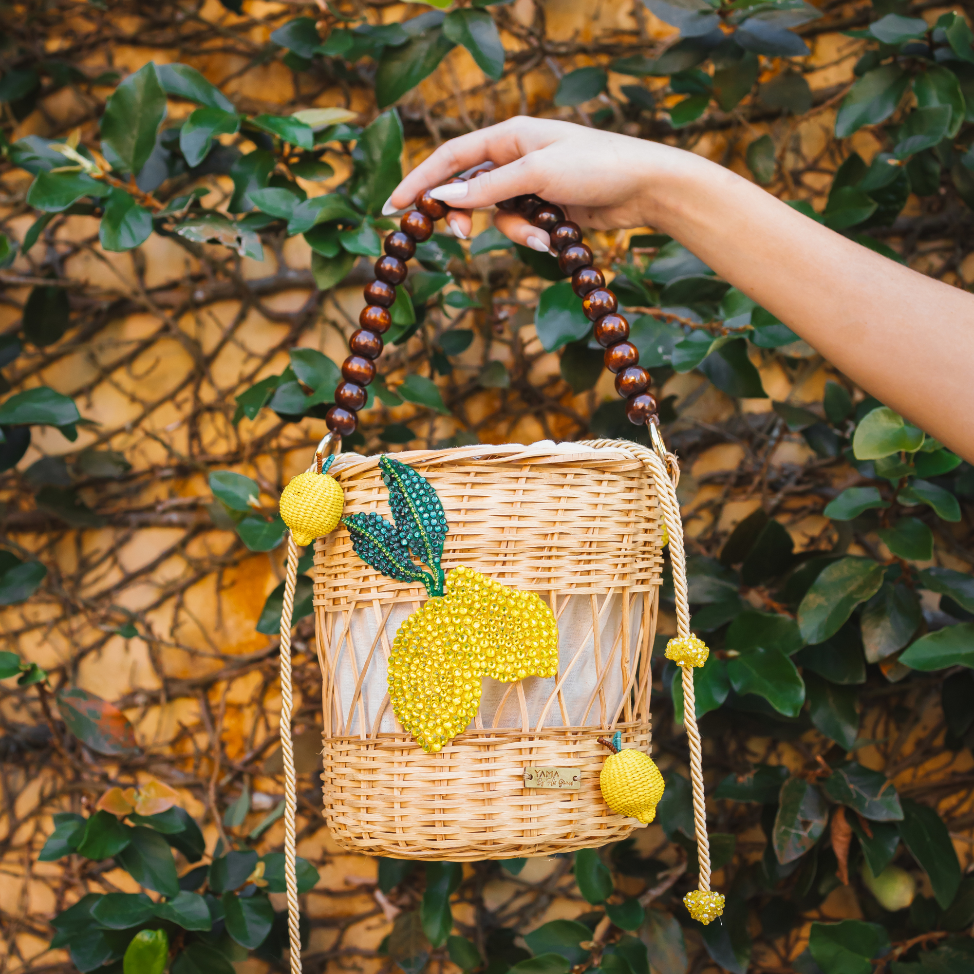 Hand holding the Lemon Delight Handbag in a leafy outdoor setting, showcasing its beaded lemon details and woven natural fiber design