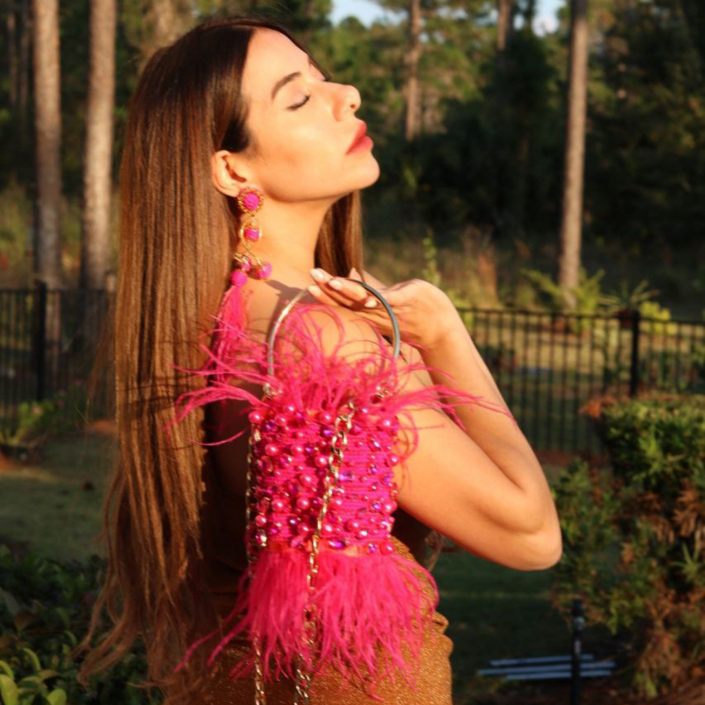 Model outdoors holding a minaudière with fuchsia feathers and gemstone details, gold chain strap visible.