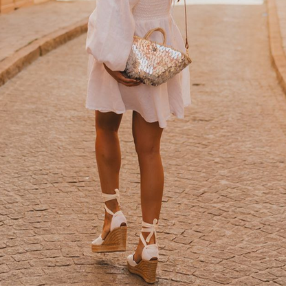 Boho-style crossbody bag with mirror shine and natural fibers, worn with wedge sandals and white dress
