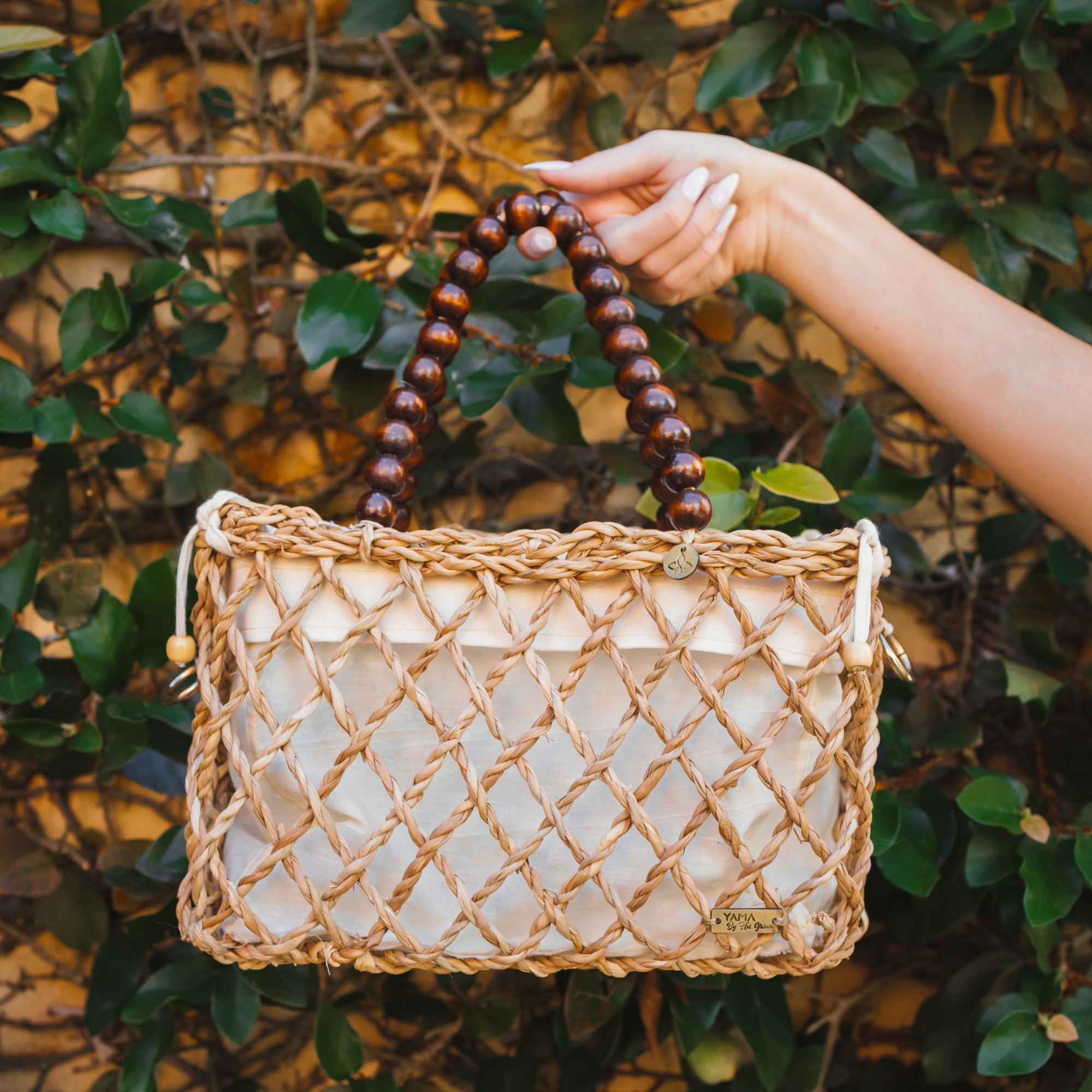 Hand holding small woven tote bag with wooden bead handles in front of green leaves