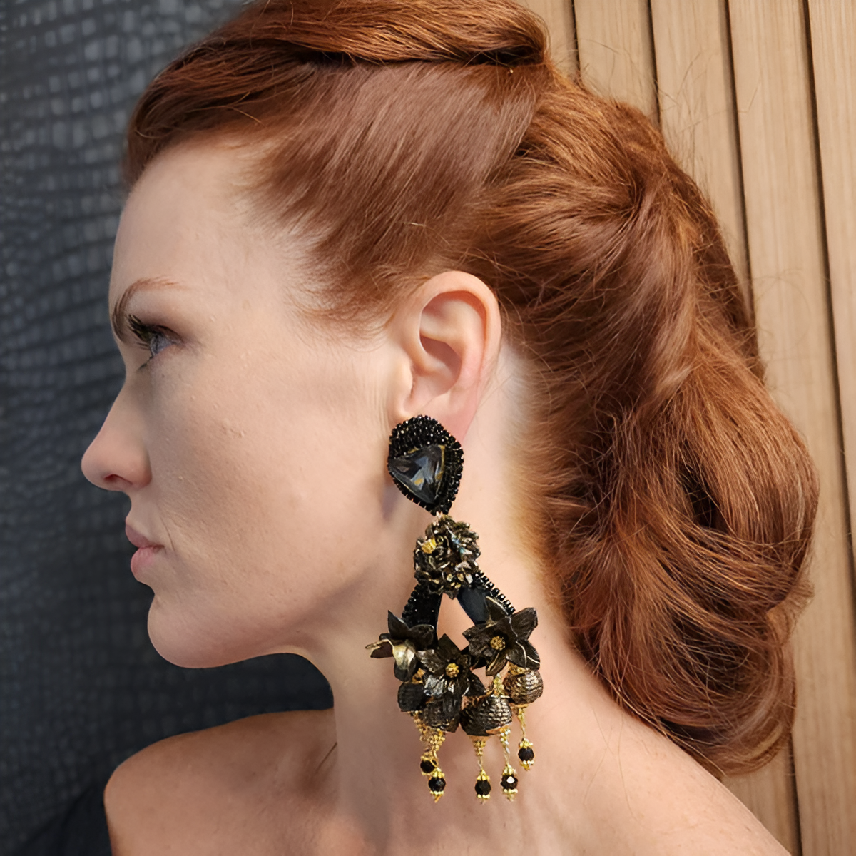 Side profile of a woman with red hair wearing a large handcrafted black and gold floral earring with crystal and beaded details.
