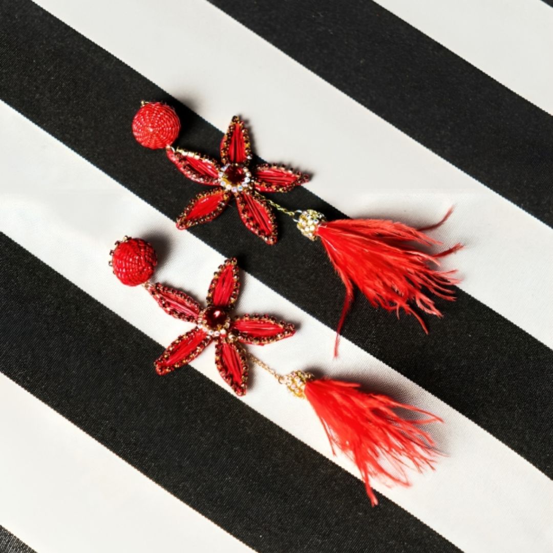 Two red floral earrings with gold and crystal details laid out on a black and white striped surface.