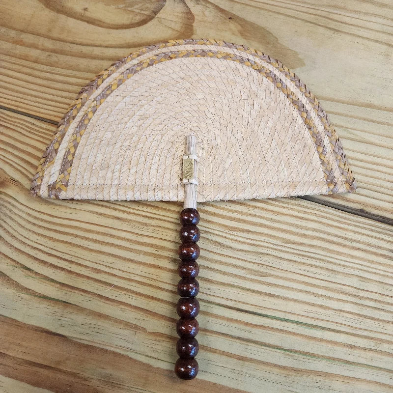 This hand fan is made by Colombian artisans using woven materials. The top of the fan features a design with alternating woven patterns of light and dark brown, creating an arched or semicircular shape