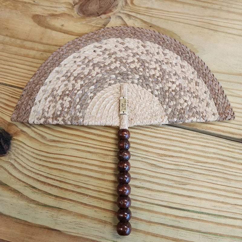 This hand fan is made by Colombian artisans using woven materials. The top of the fan features a design with alternating woven patterns of light and dark brown, creating an arched or semicircular shape