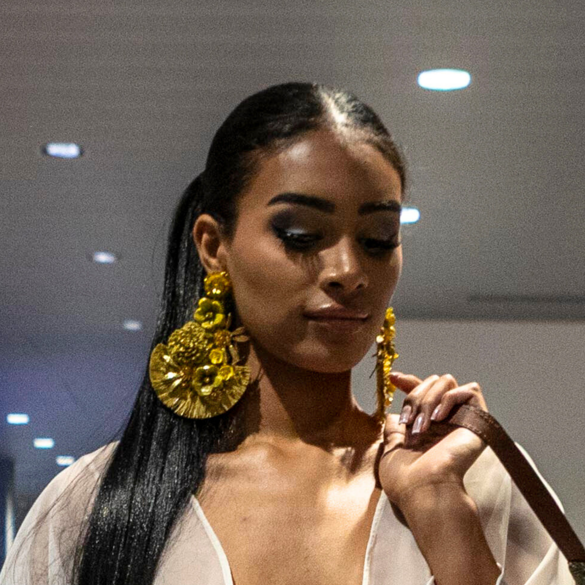 Elegant woman with long straight hair wearing large gold raffia earrings featuring floral and leaf embellishments, walking indoors at a formal event.