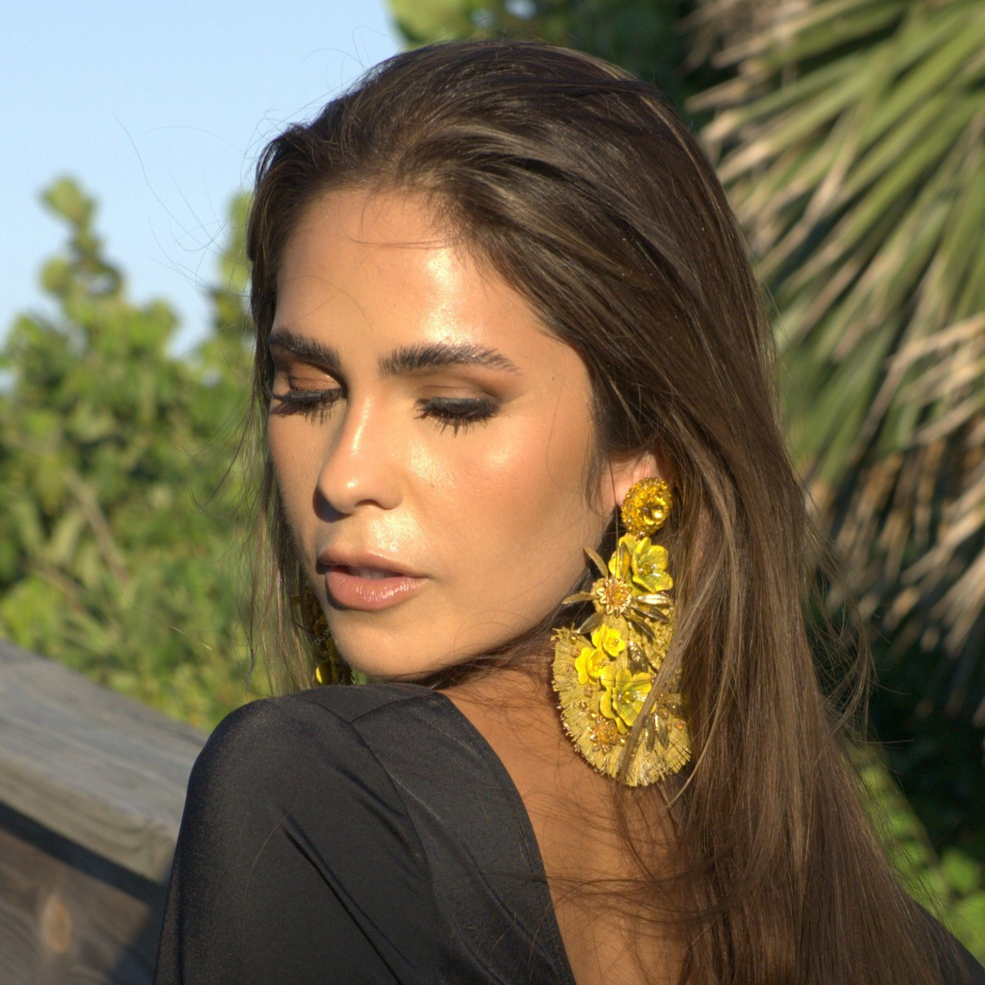 Woman wearing golden floral statement earrings with raffia detailing, captured in soft natural light against a lush outdoor backdrop.