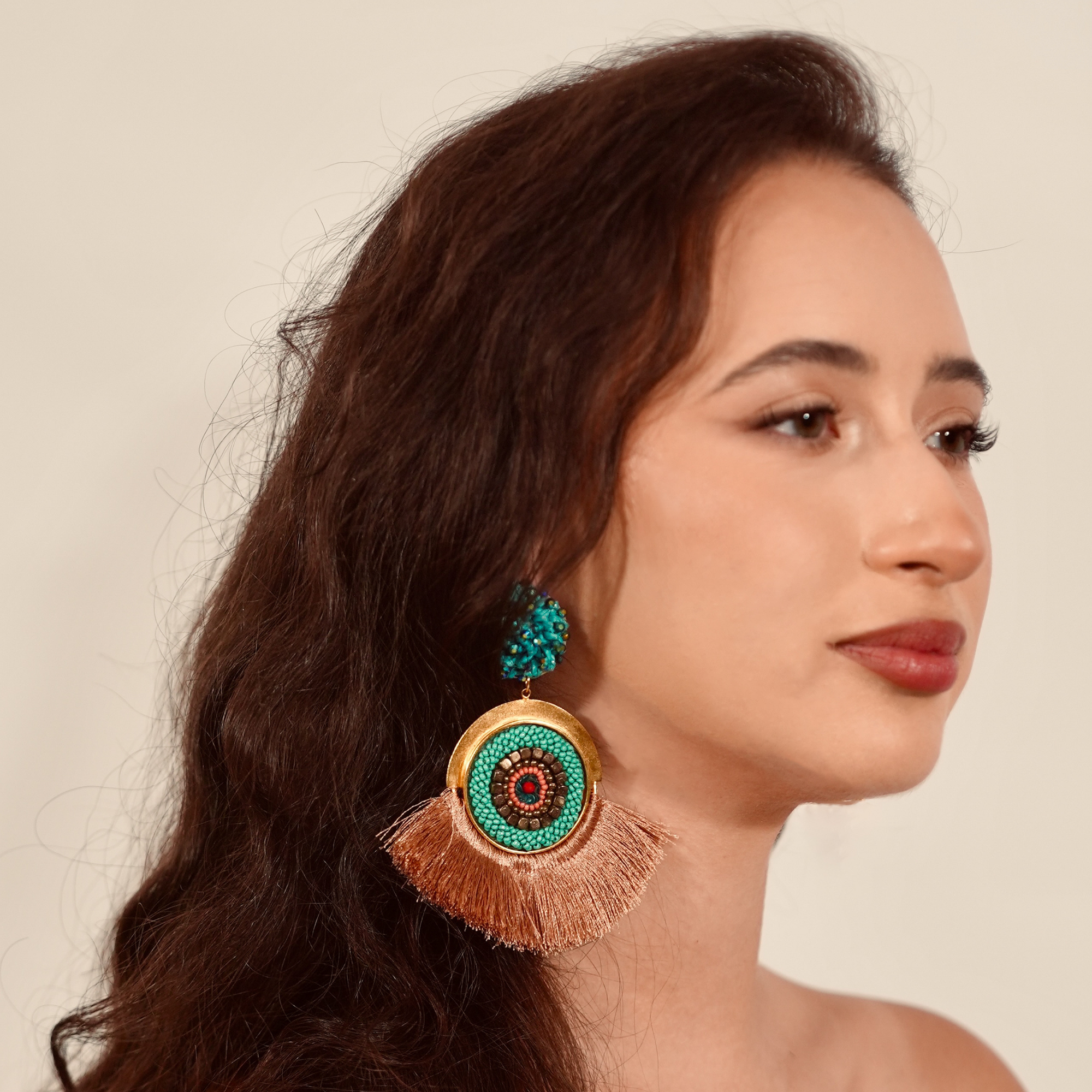 Woman wearing a pair of earrings with turquoise and gold design on a beige background