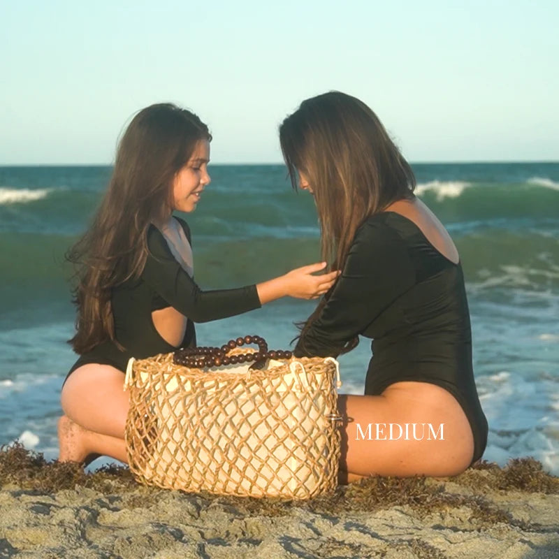 Mother and daughter sitting on the beach with a medium-size woven tote bag between them