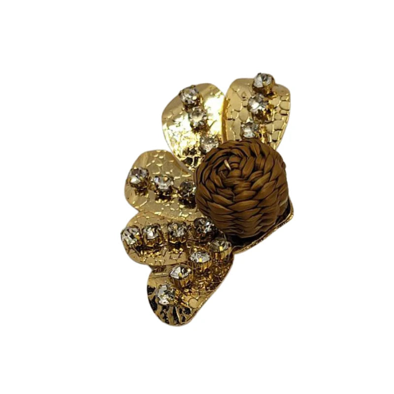 This stunning brooch showcases an elegant floral design, featuring shimmering golden petals with intricate hammered textures and dazzling crystal accents. At its center lies a beautifully woven banana fiber sphere, adding a natural, artisanal touch that contrasts gracefully with the metallic brilliance.