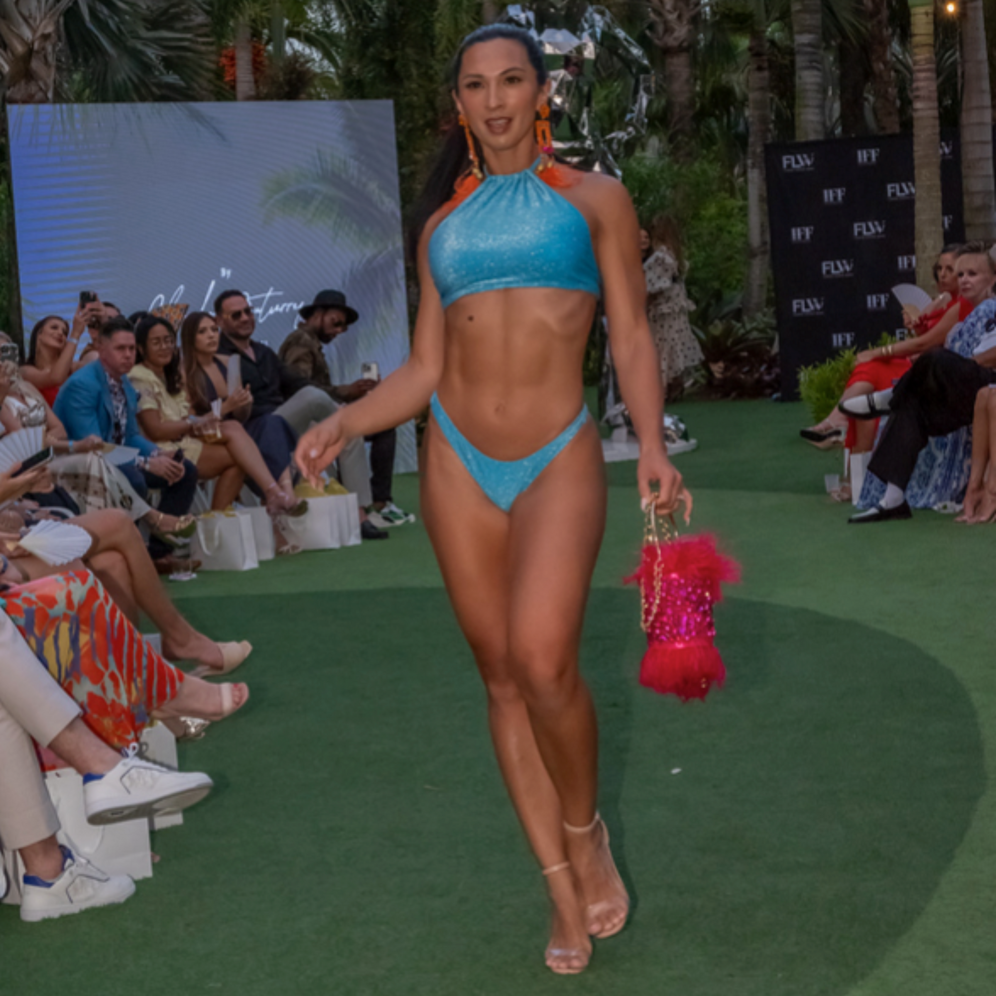 Runway model in a turquoise bikini carrying the fuchsia feather minaudière with gold chain on a fashion show catwalk.